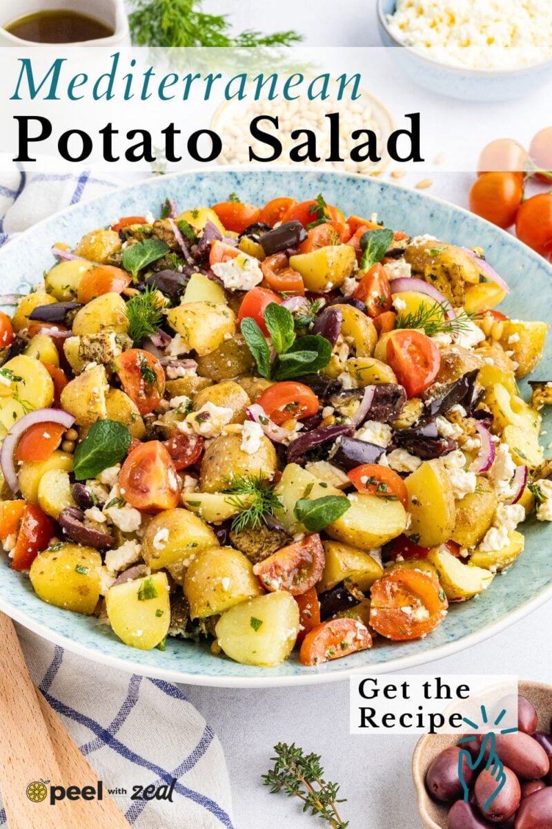 A bowl of Greek potato salad with cherry tomatoes, olives, red onions, feta cheese, and herbs on a table with eggs and extra feta in the background.