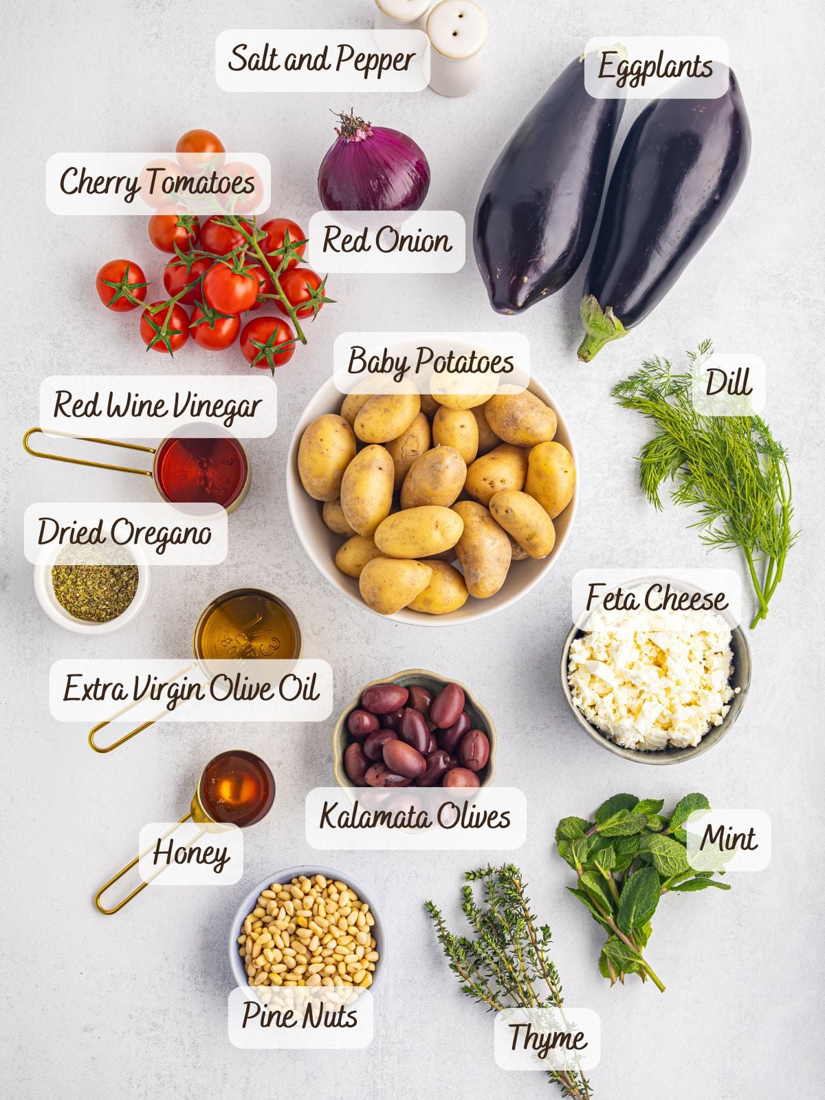 Various ingredients for a Greek potato salad are arranged on a white surface, including eggplants, baby potatoes, cherry tomatoes, Kalamata olives, feta cheese, herbs, olive oil, and seasonings.