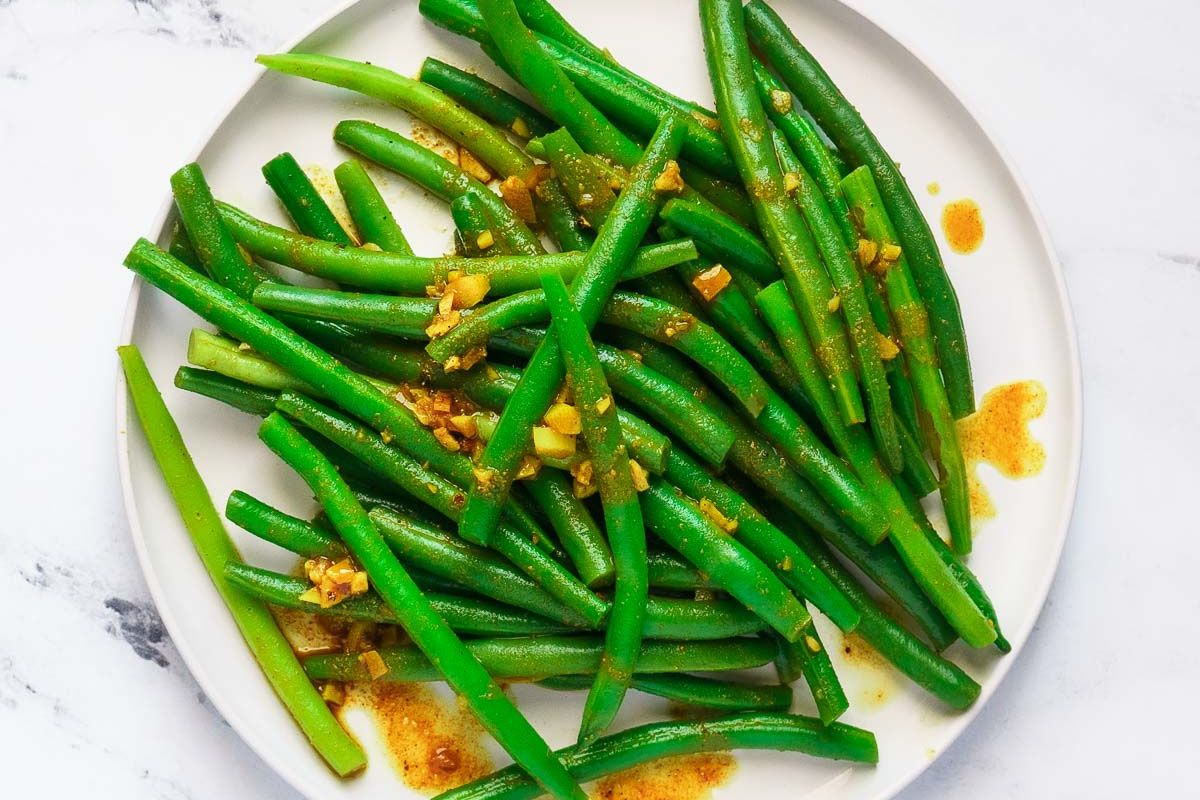 A white plate with cooked green beans topped with a chopped garlic and spice mixture, with some seasoning sauce visible on the plate.