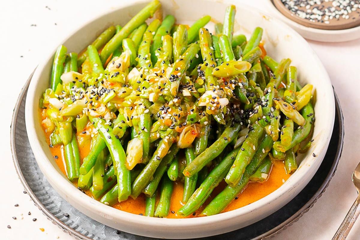 A bowl of green beans in sauce, topped with chopped scallions, sesame seeds, and a sprinkle of seasoning.