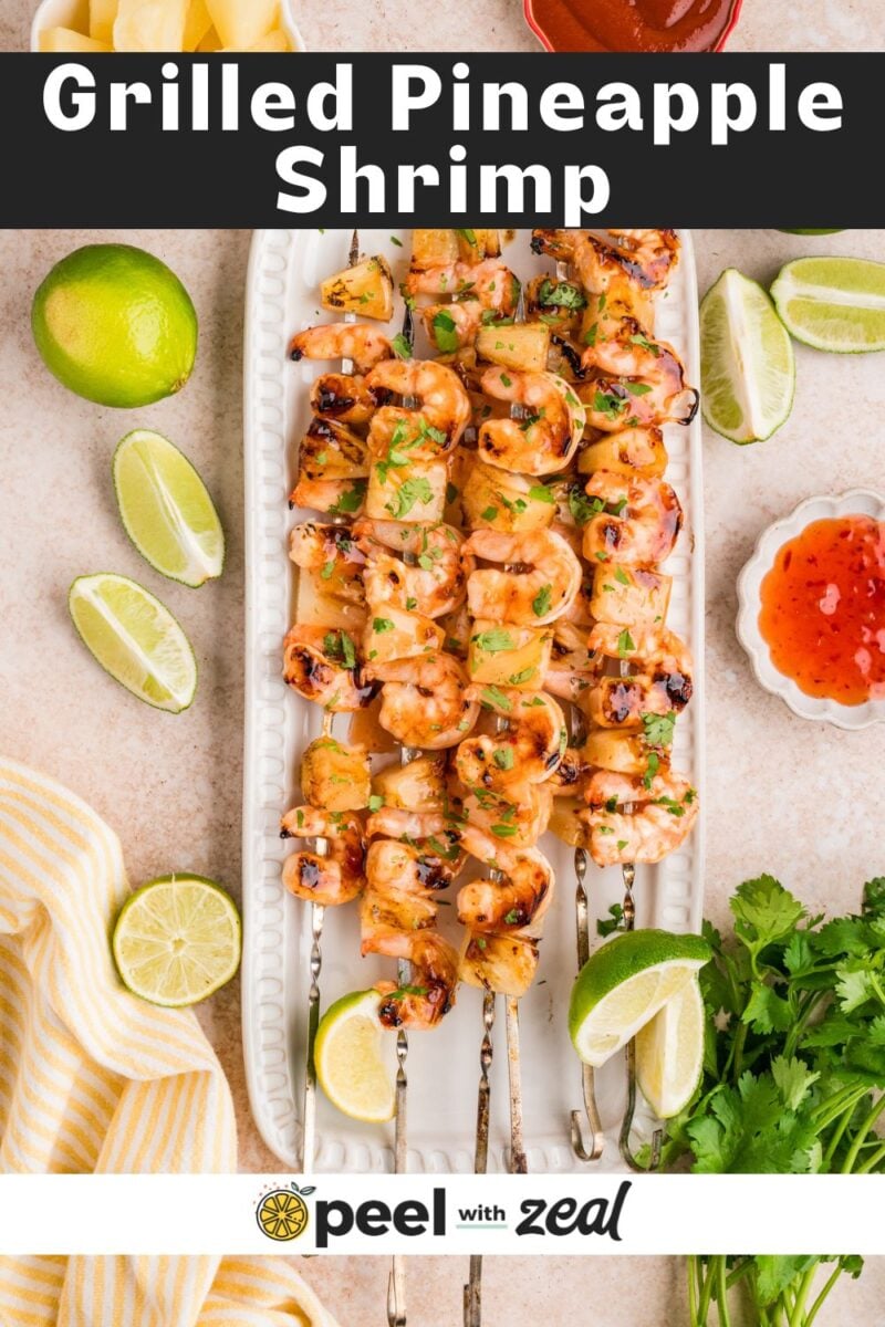 A platter of grilled pineapple shrimp skewers garnished with cilantro, surrounded by lime wedges, cilantro, a bowl of sweet chili sauce, and a yellow-striped towel.