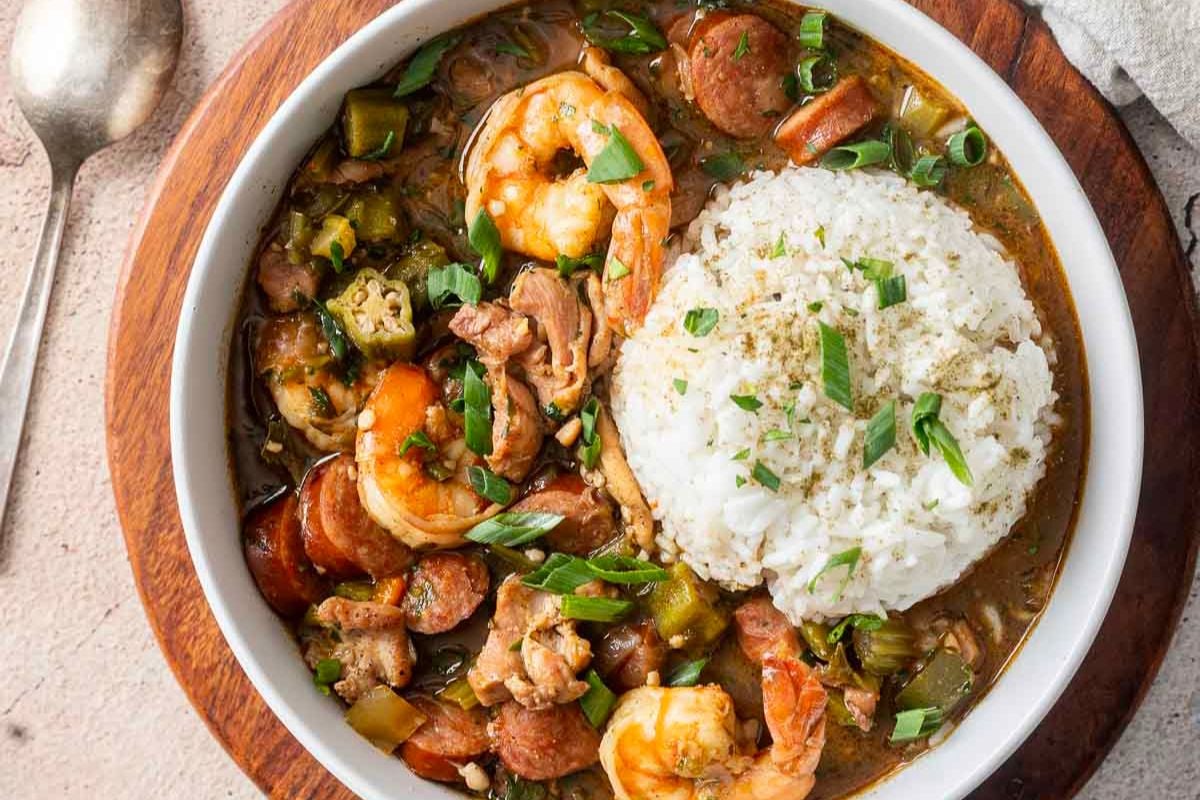 A bowl of gumbo with shrimp, sausage, and vegetables, served with a mound of white rice and garnished with chopped green onions.