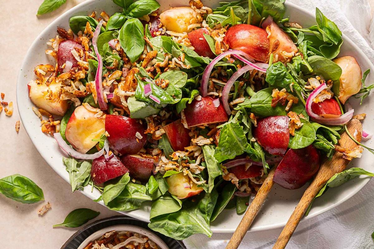 A bowl of fresh spinach salad with sliced nectarines, red onions, crispy fried onions, and basil leaves, served with wooden salad utensils.