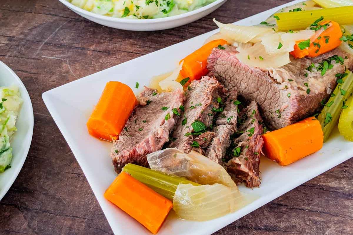 A plate of comforting beef pot roast with cooked carrots, celery, and onions, garnished with chopped parsley. Mashed potatoes are visible in the background