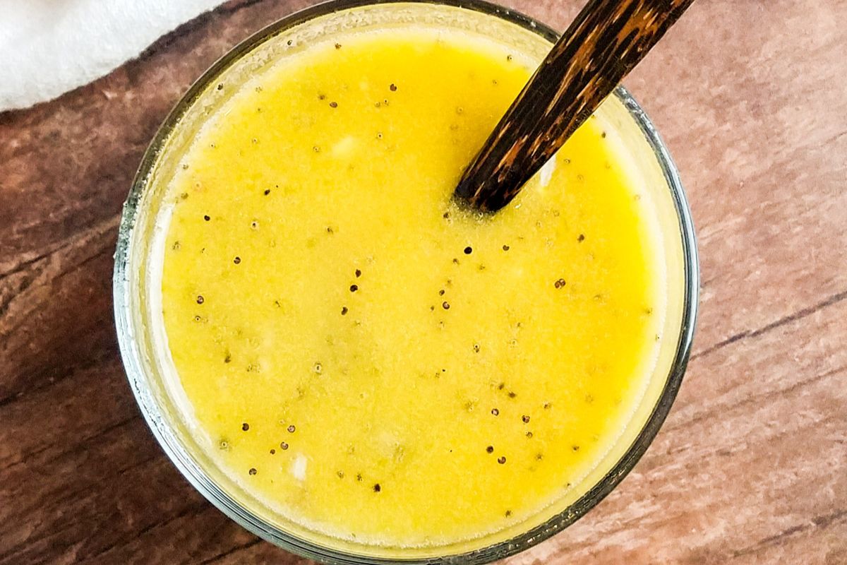 A glass bowl filled with yellow salad dressing containing visible spices, with a dark wooden spoon resting inside, on a wooden surface.