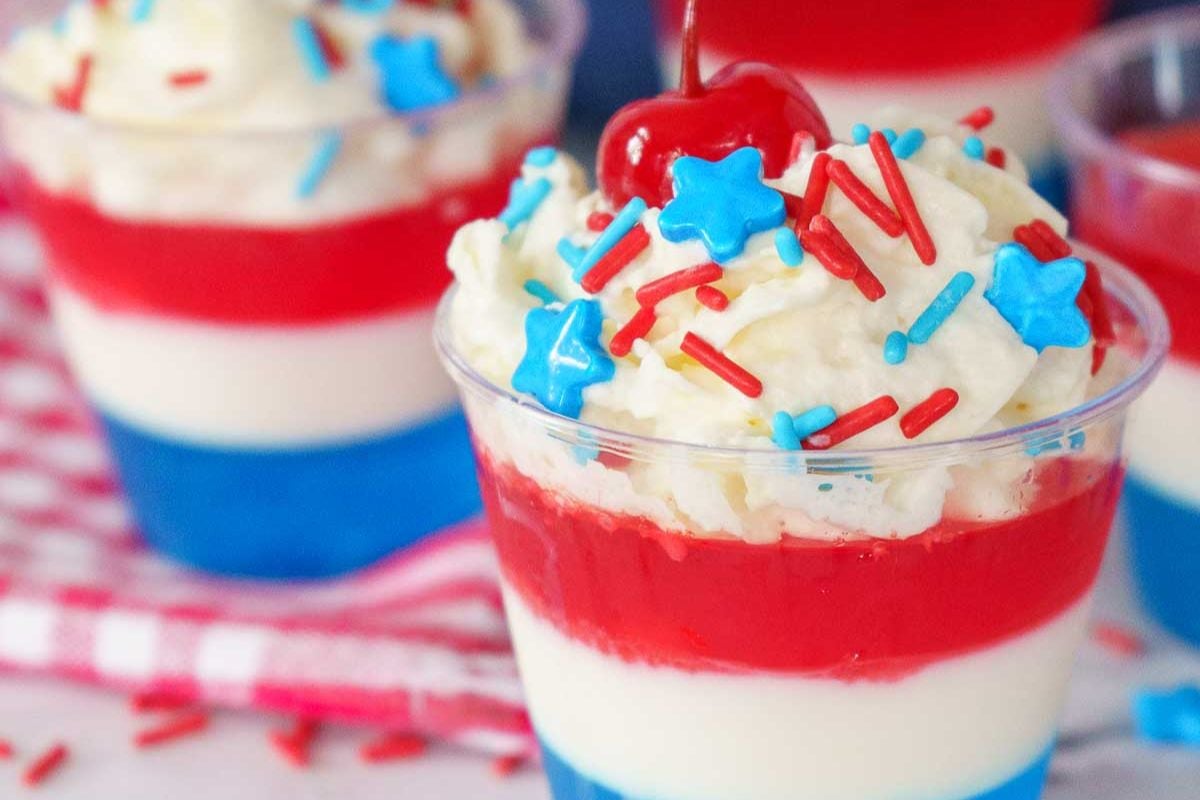 A layered dessert in a clear cup with blue, white, and red layers, topped with whipped cream, star-shaped sprinkles, red sprinkles, and a cherry.