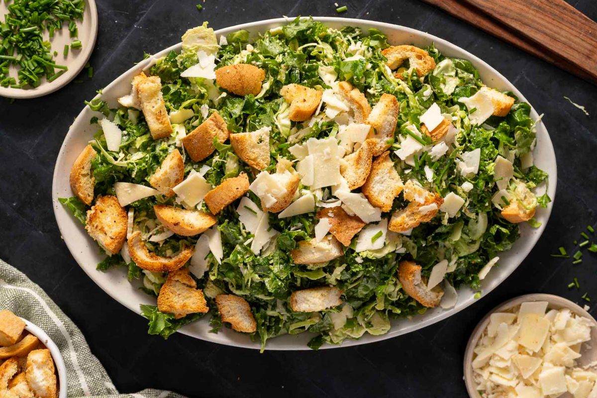 A large oval platter of Caesar salad topped with croutons and parmesan cheese shavings, placed on a dark surface with small bowls of toppings nearby.