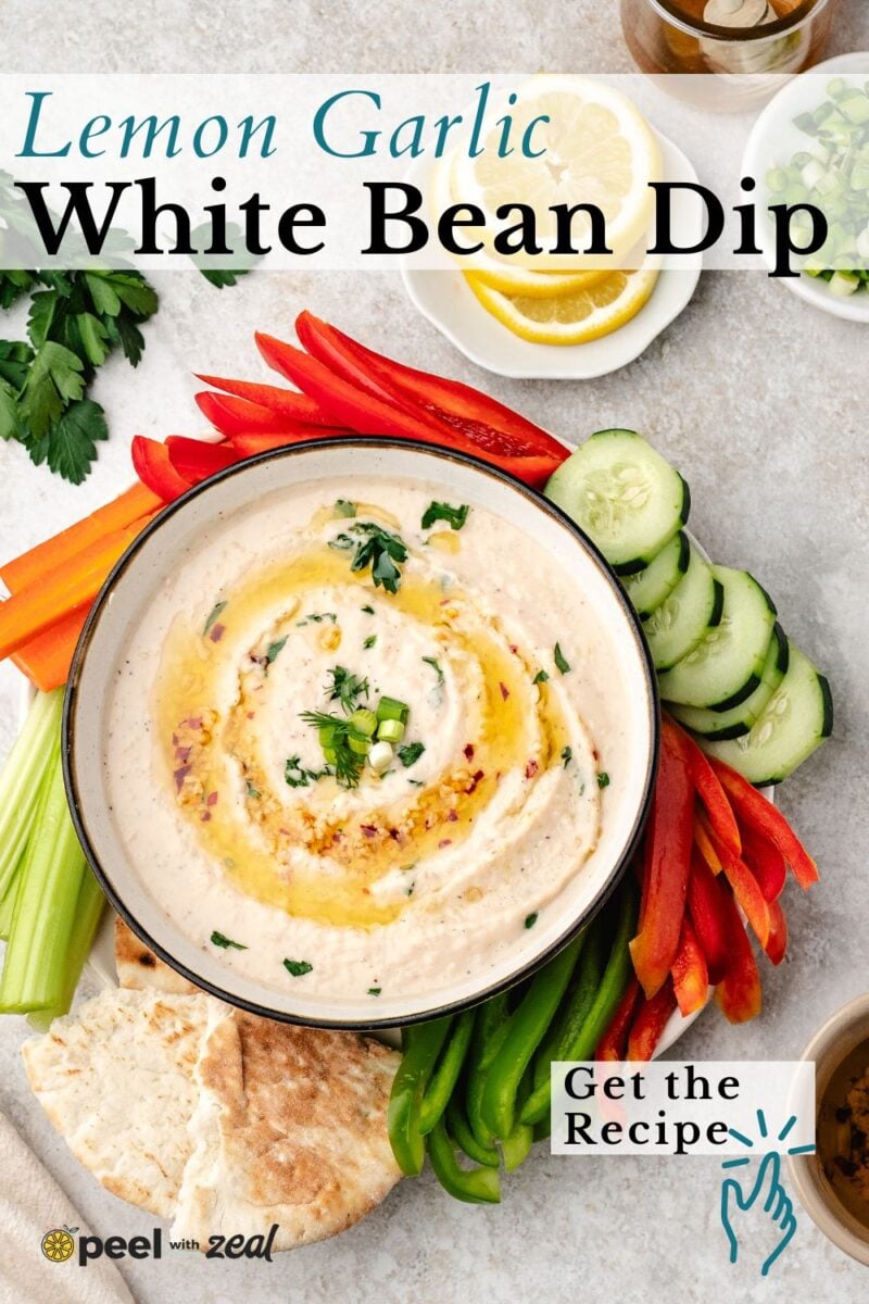Bowl of creamy White Bean Dip with lemon and garlic, garnished with fresh herbs and olive oil, surrounded by sliced vegetables, pita bread, and lemon slices on a light surface.