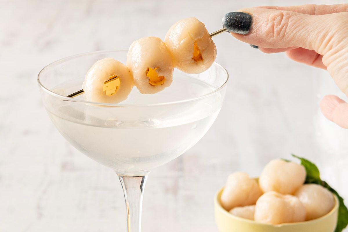 A hand holds a skewer of lychee fruits over a clear cocktail in a stemmed glass; a small bowl of lychees sits nearby.