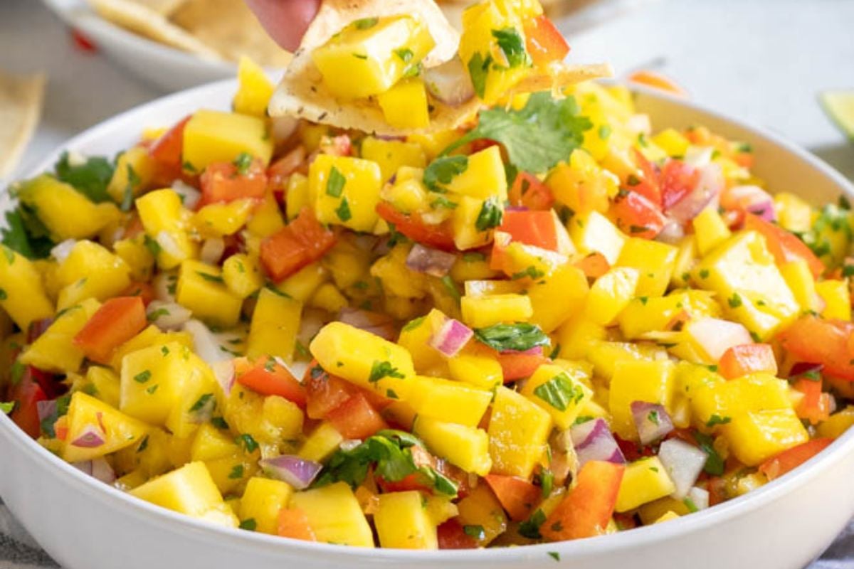A bowl of mango salsa with diced mango, red bell pepper, red onion, cilantro, and a hand dipping a tortilla chip into the salsa.