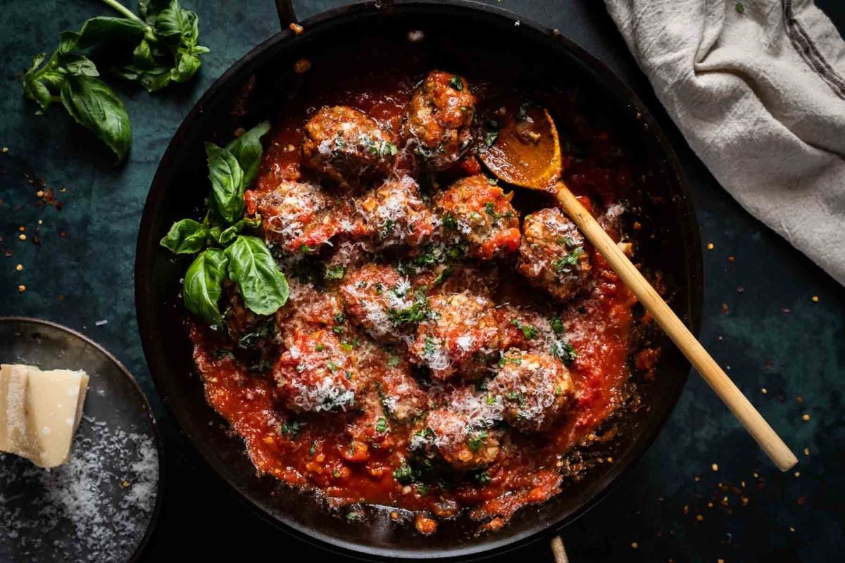 Meatballs covered in tomato sauce and grated cheese in a skillet, garnished with basil, with a wooden spoon and a cloth nearby.