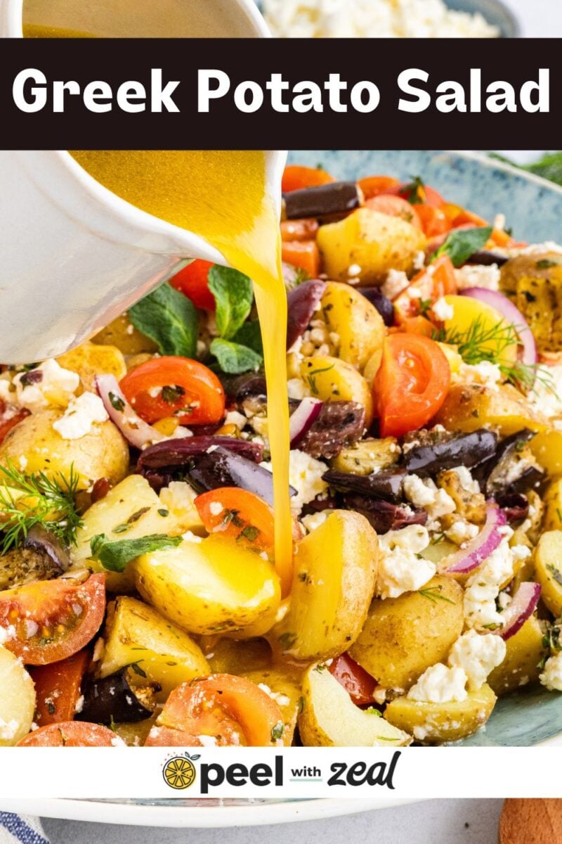 A bowl of Greek potato salad with tomatoes, olives, red onions, feta cheese, and herbs, as a tangy dressing is poured on top.