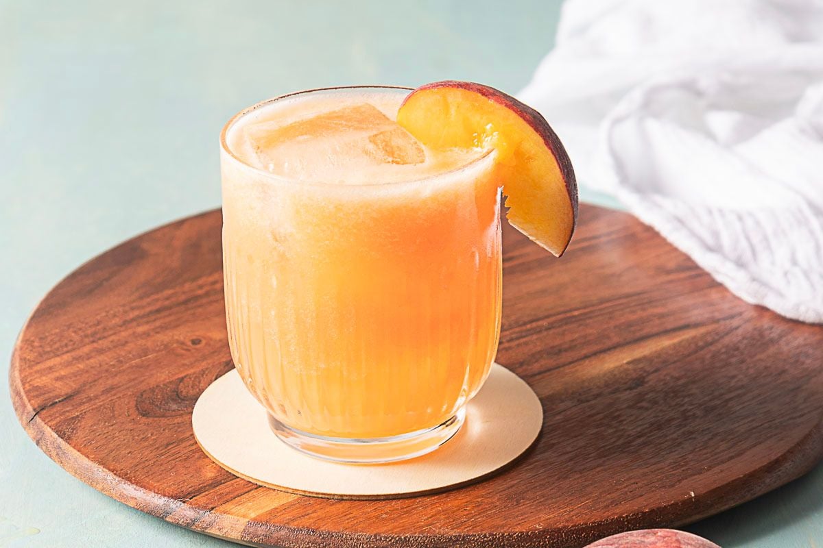 A glass of orange-colored beverage with ice and a peach slice garnish sits on a round wooden board with a white napkin in the background.