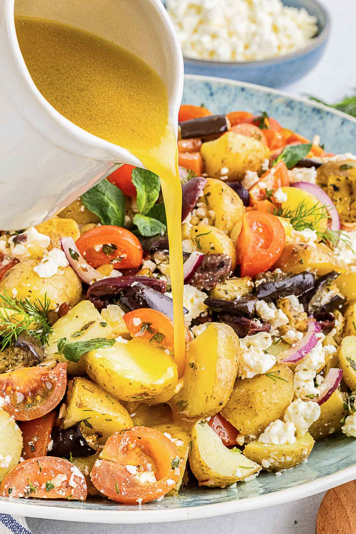 A close-up of a salad with potatoes, cherry tomatoes, olives, red onions, and feta cheese as a dressing is poured over it from a white pitcher.