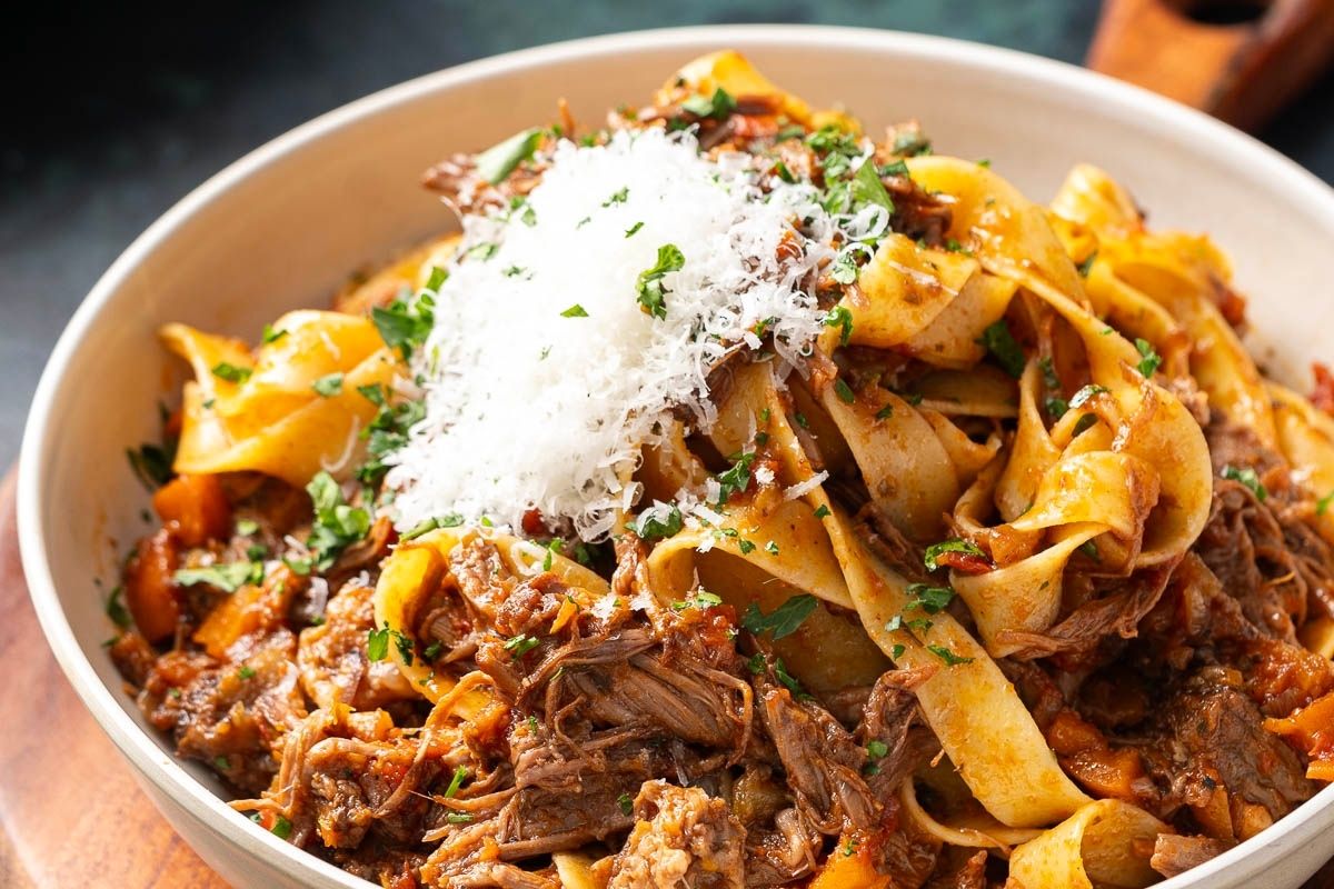 A bowl of pappardelle pasta topped with shredded meat ragu, grated cheese, and chopped parsley.