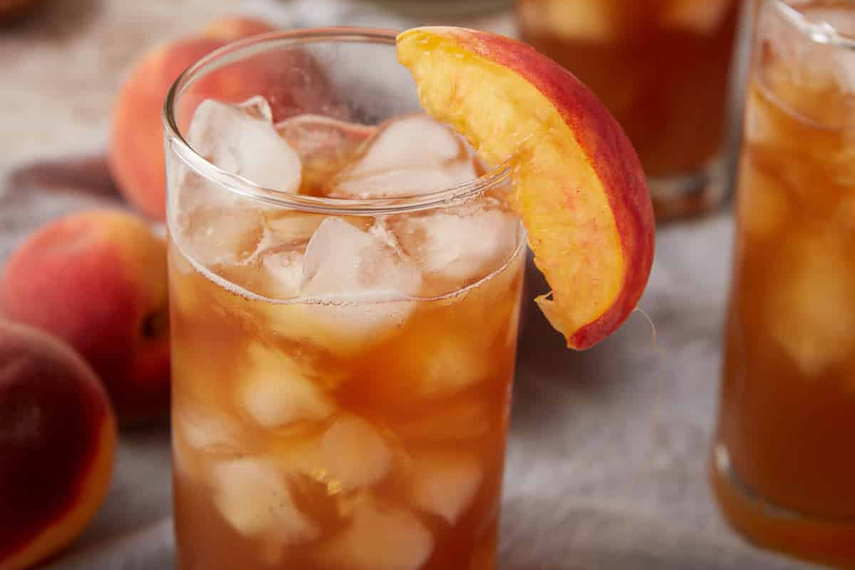 A glass of iced tea with ice cubes, garnished with a peach slice, sits on a table with whole peaches in the background&mdash;a perfect choice for refreshing beverages and classic porch drinks.