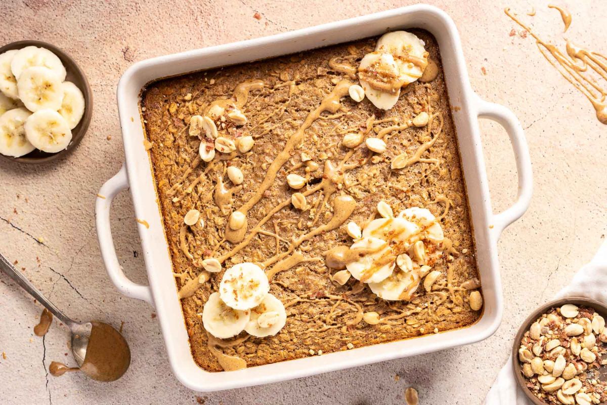A baked oatmeal dish topped with banana slices, peanut butter drizzle, and chopped nuts sits in a white square baking pan on a light-colored surface.