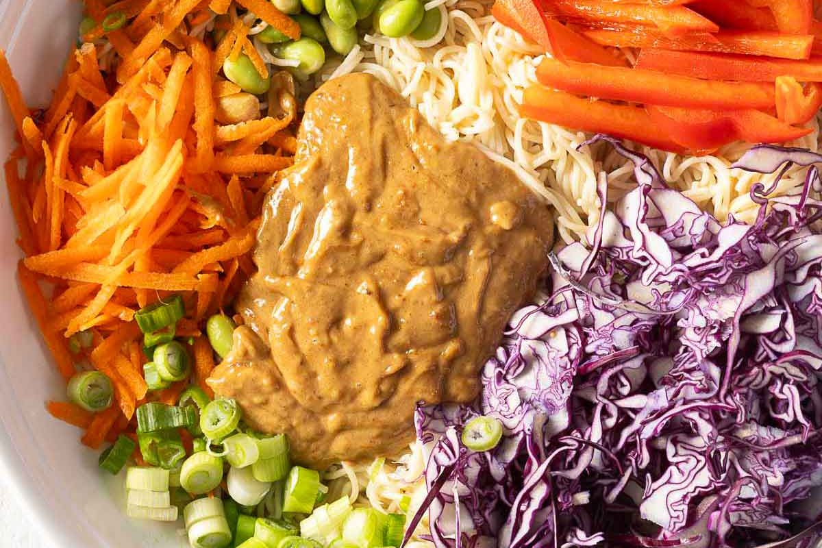 A bowl containing shredded carrots, sliced green onions, chopped cabbage, red bell pepper strips, noodles, and a large dollop of peanut sauce in the center.
