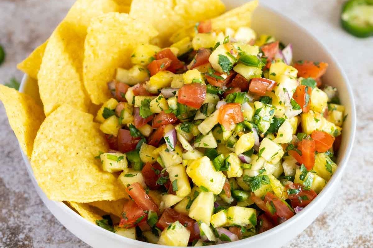 A bowl of pineapple salsa with chopped tomatoes, onions, cilantro, and jalapeños, served with tortilla chips on the side.