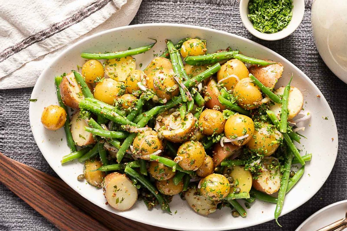 A platter of potato and green bean salad with herbs, thinly sliced onions, and seasoning, served on a white oval dish. A small bowl of chopped herbs is nearby.