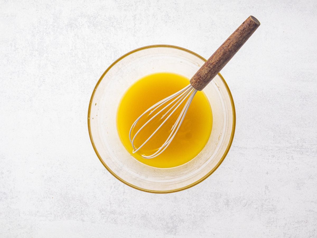 A glass bowl with a yellow liquid and a whisk with a wooden handle on a light-colored surface.