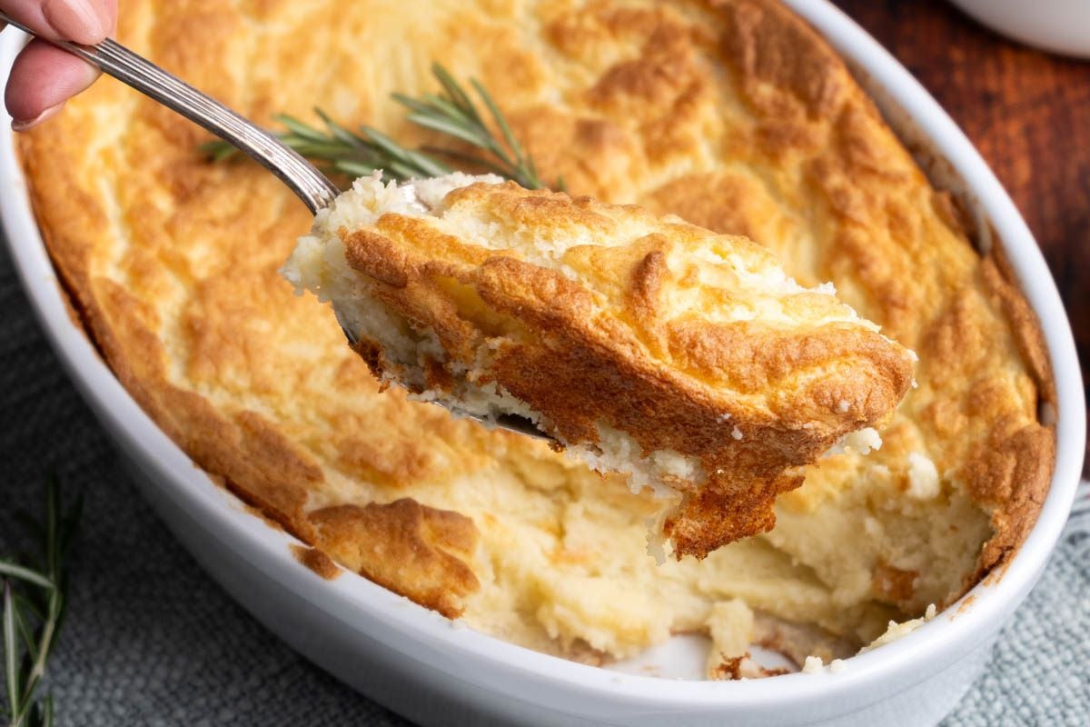 A spoon lifts a serving of golden-brown mashed potato casserole from a white baking dish, garnished with a sprig of rosemary.