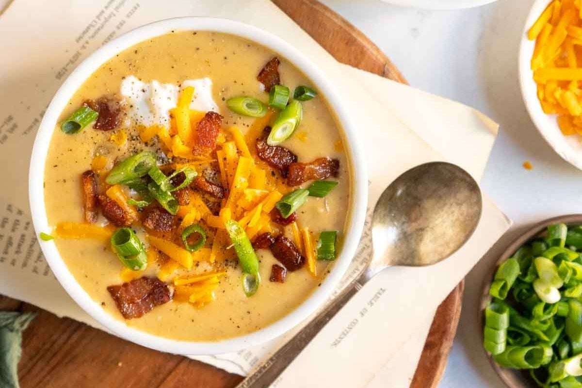 A bowl of creamy soup topped with shredded cheddar cheese, bacon pieces, and chopped green onions, with a spoon and green onions on the side.
