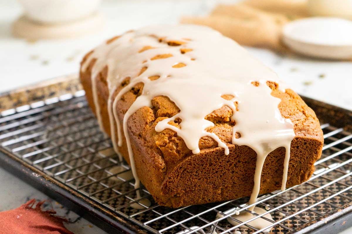 A loaf of bread with white icing drizzled on top sits on a wire rack over a baking sheet.