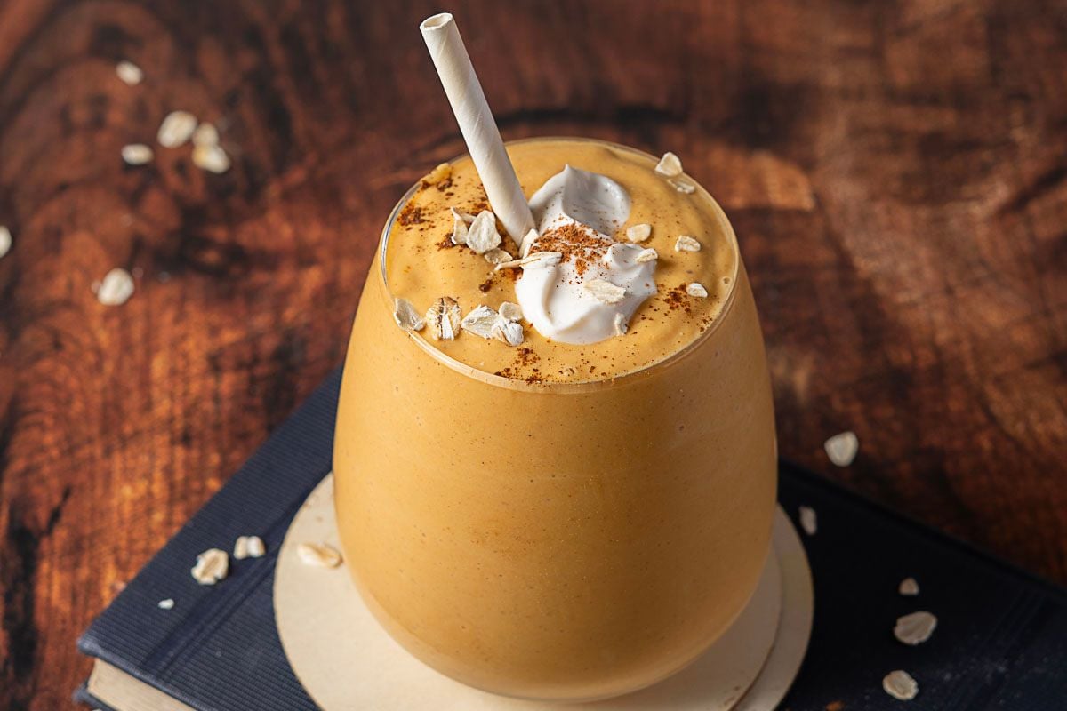 A glass of creamy orange smoothie topped with whipped cream, oats, and a sprinkle of cinnamon, with a paper straw on a wooden table.