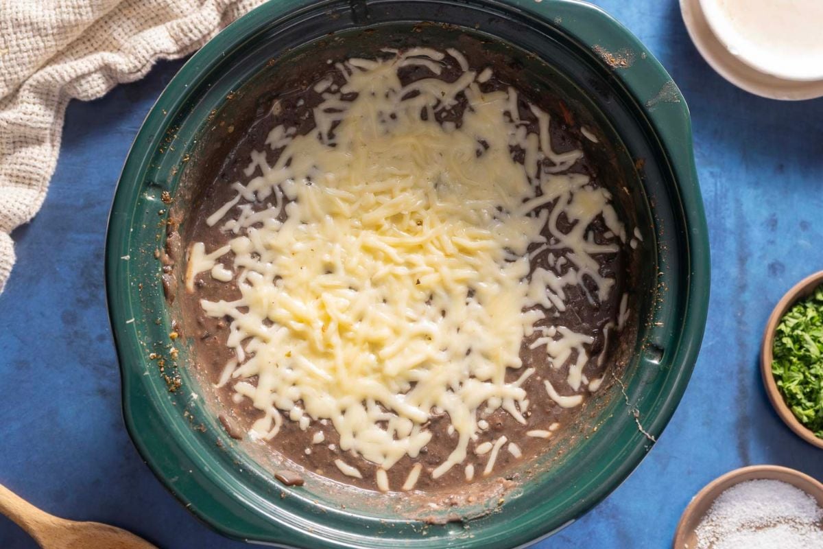 A slow cooker filled with melted shredded cheese on top of black refried beans, surrounded by various small dishes on a blue surface.