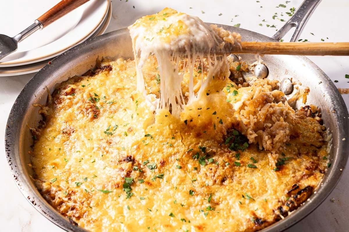 A skillet filled with cheesy, baked rice being scooped up with a wooden spoon; melted cheese stretches from the pan.