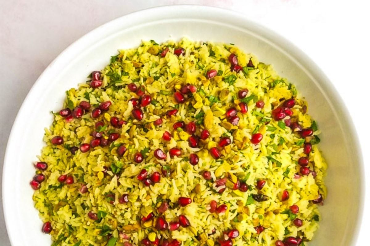 A white plate filled with yellow rice mixed with herbs and topped with pomegranate seeds and pistachios.