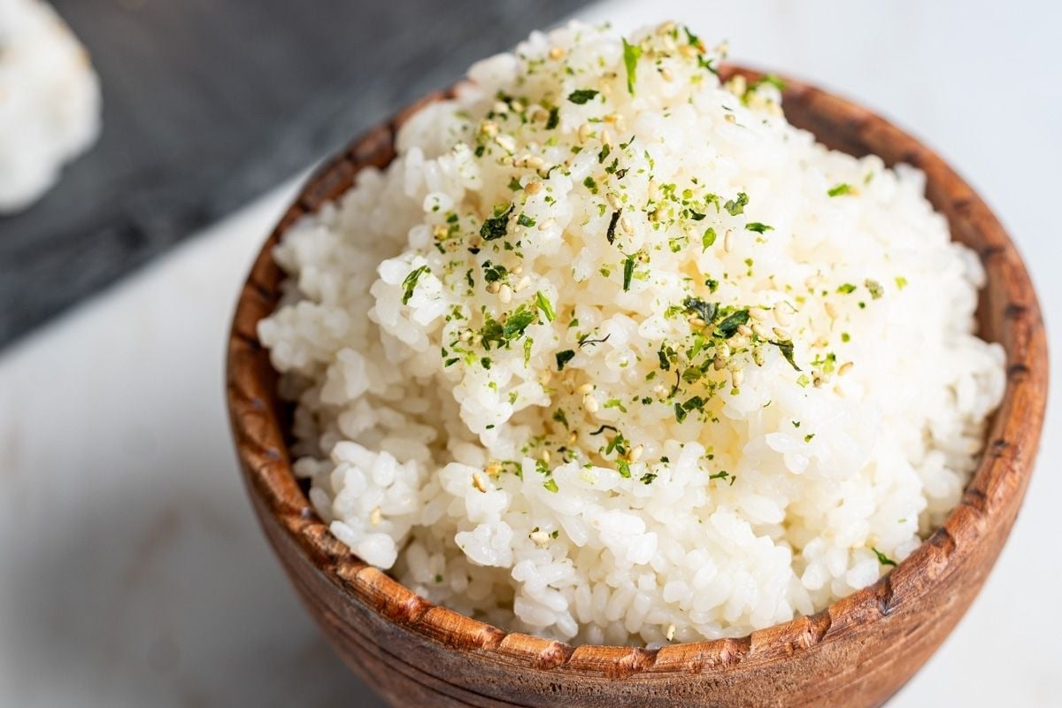 A wooden bowl filled with cooked white rice, topped with a sprinkle of green herbs or seasoning.