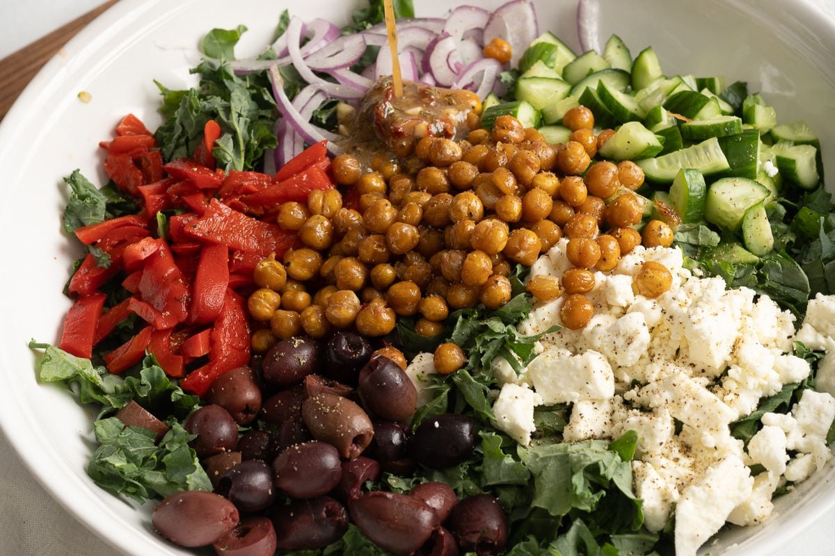 A bowl of salad with kale, roasted chickpeas, feta cheese, Kalamata olives, red peppers, cucumber, red onion, and dressing being poured on top.