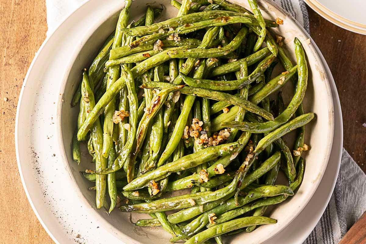 A bowl of cooked green beans garnished with minced garlic, served on a white plate.