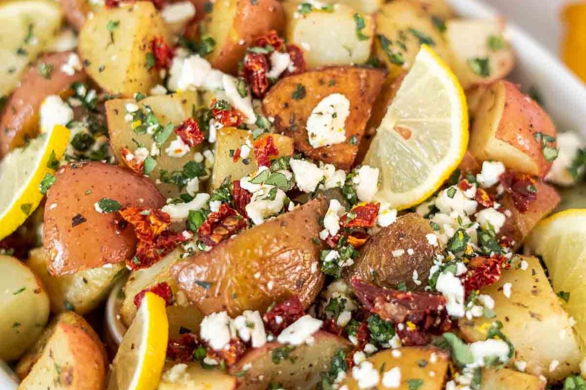 A bowl of roasted potatoes garnished with lemon wedges, crumbled cheese, chopped herbs, and pieces of sun-dried tomato.