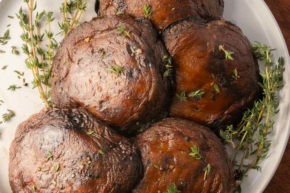 Five roasted portobello mushroom caps on a white plate, garnished with sprigs of fresh thyme.