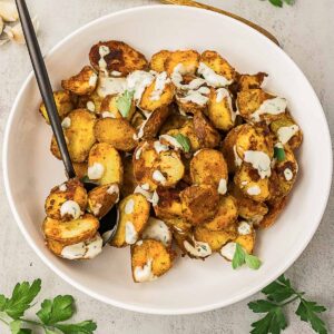 A white bowl filled with roasted potato slices topped with a creamy sauce and garnished with parsley, with a black serving spoon on the side.
