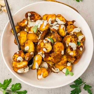 A white bowl filled with roasted potato slices topped with a creamy sauce and garnished with parsley, with a black serving spoon on the side.