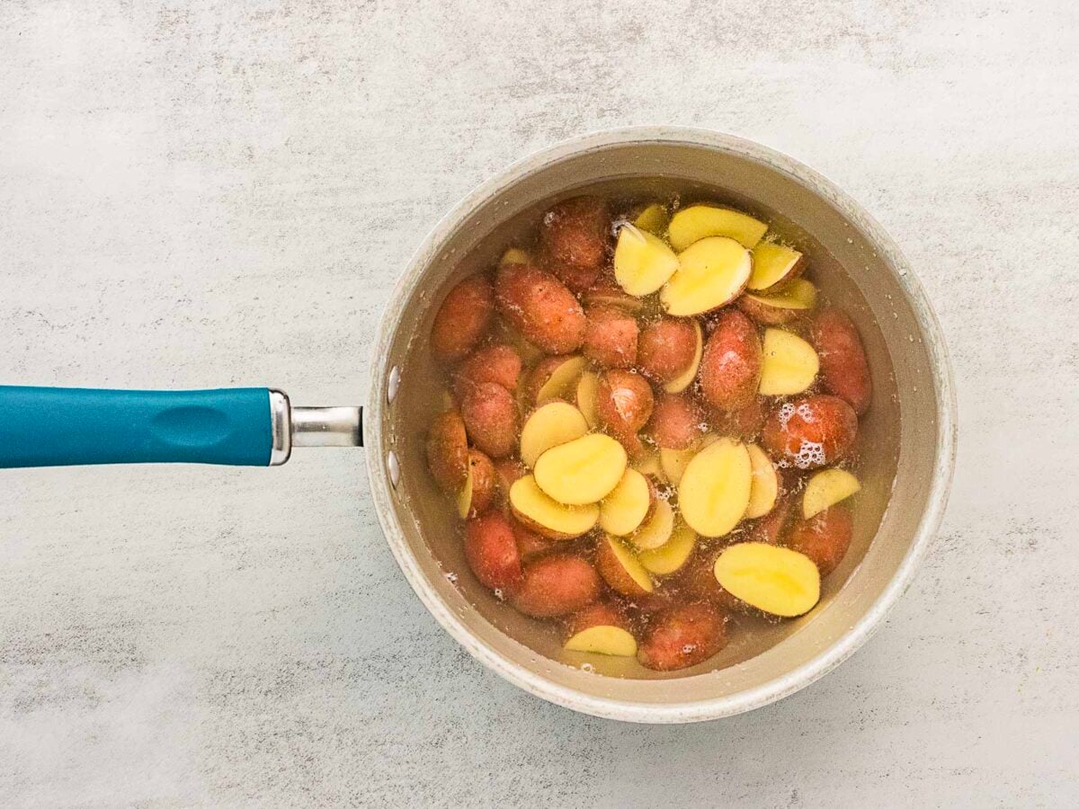 A saucepan filled with sliced red potatoes submerged in water, placed on a light-colored surface.
