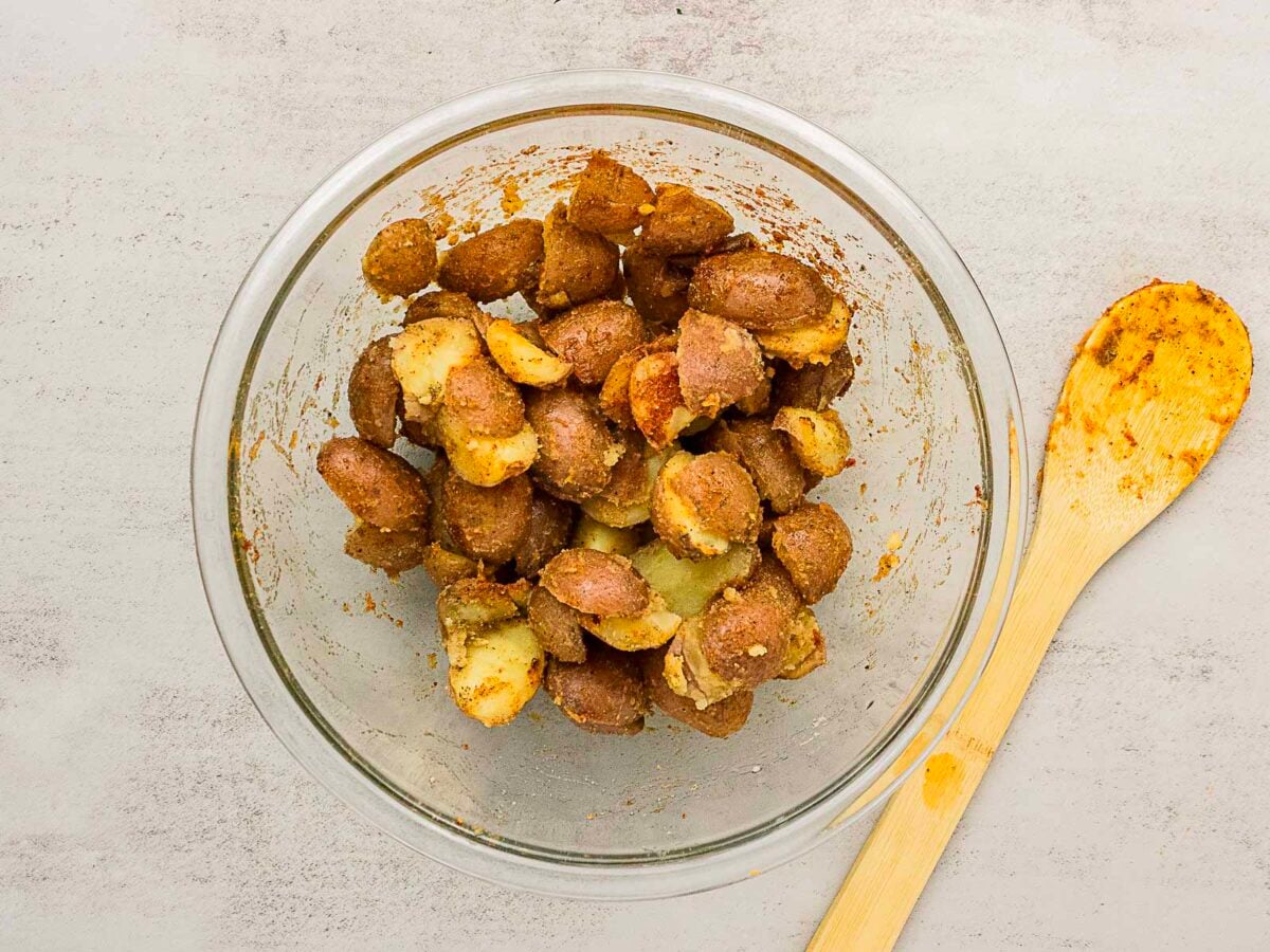 Glass bowl containing seasoned, halved baby potatoes next to a wooden spoon with seasoning residue on a light-colored surface.