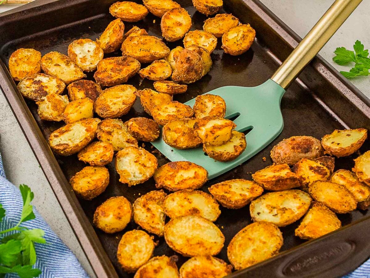Roasted, seasoned potato halves are spread on a baking sheet, with a spatula lifting some of them. Fresh parsley and a striped towel are nearby.