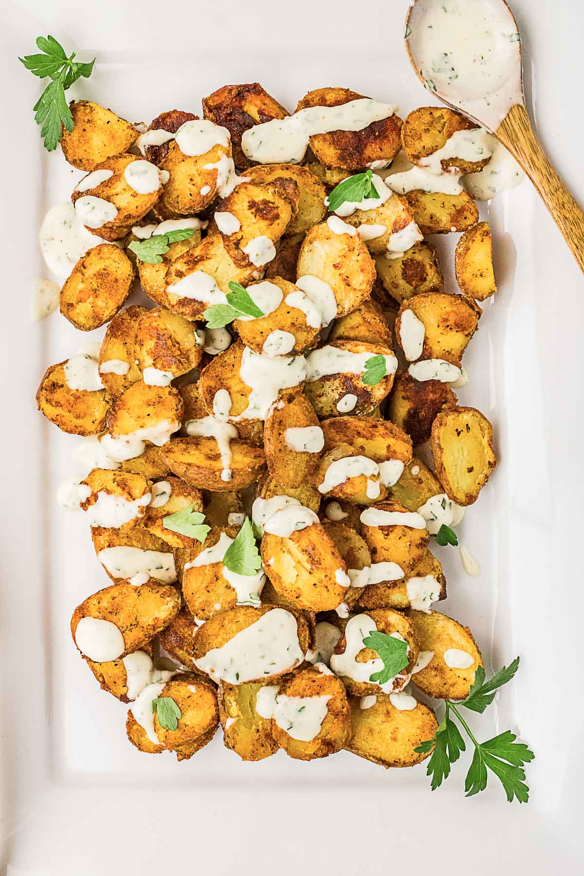 Roasted potato pieces topped with a creamy white sauce and garnished with fresh parsley, served on a rectangular white platter with a wooden spoon.