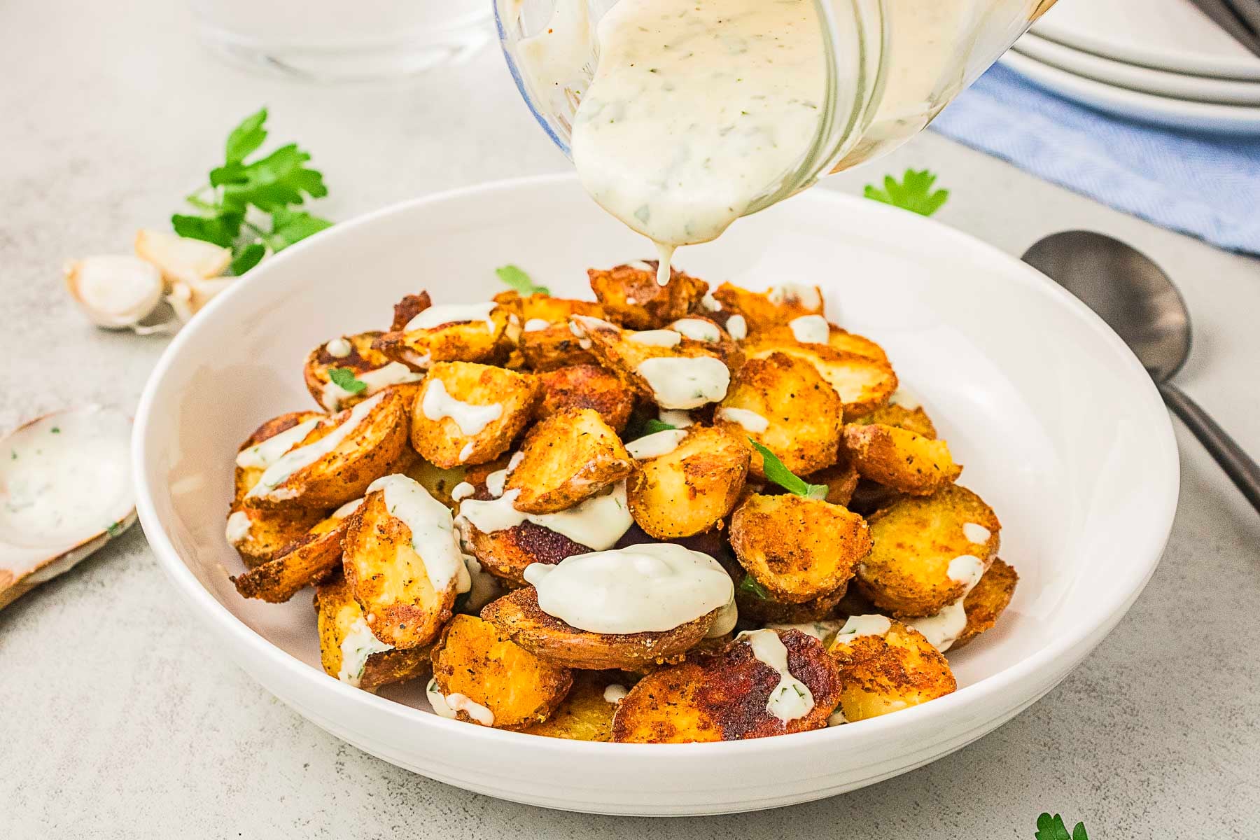 A bowl of roasted potato slices being drizzled with creamy white sauce, garnished with parsley, on a light-colored table.