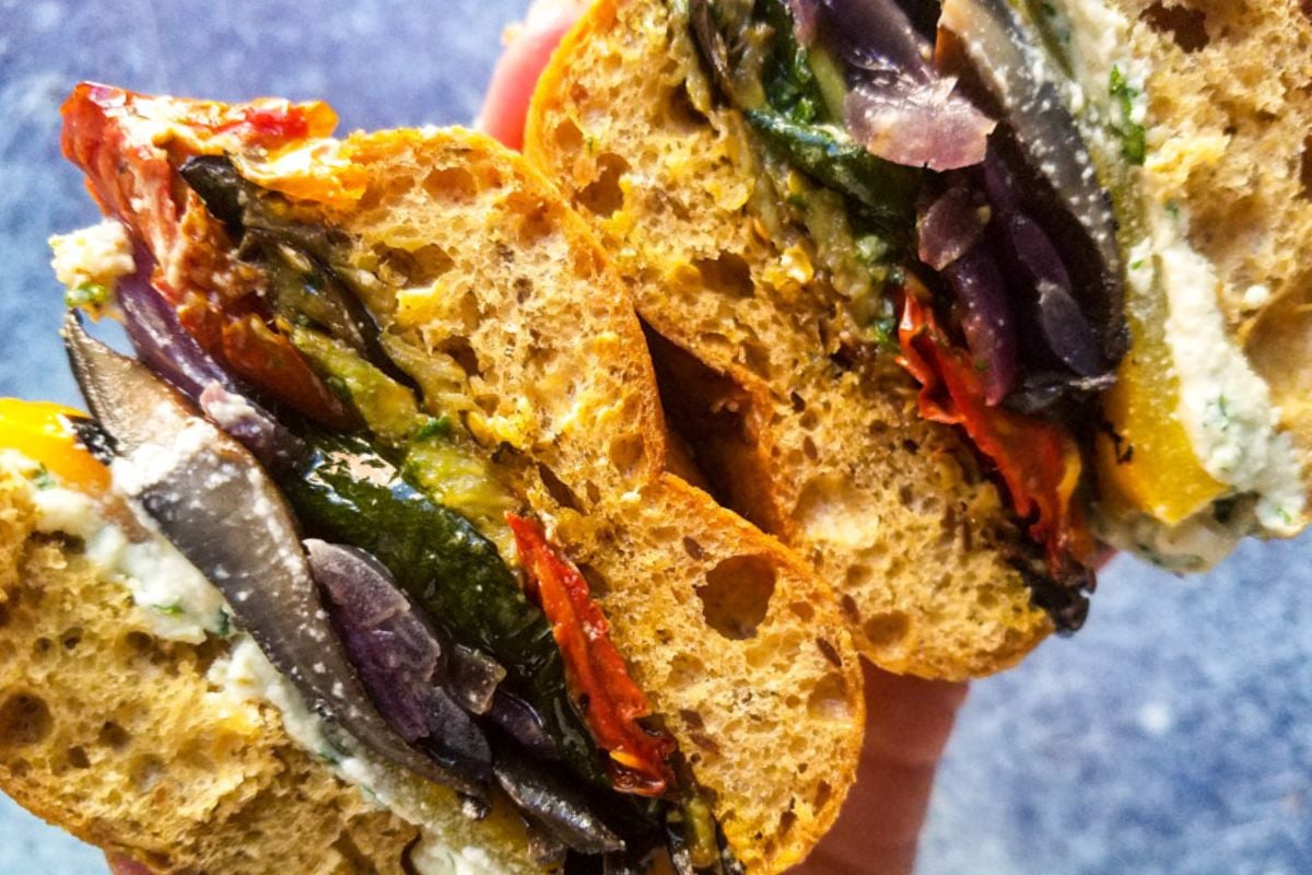 A close-up of a halved sandwich filled with roasted vegetables and a white cheese spread on brown bread, held up against a blue background.