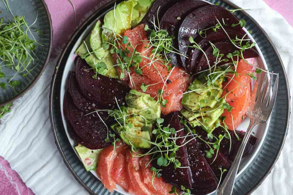 A plate of sliced beets, avocado, and grapefruit, topped with microgreens, with a fork on the side.