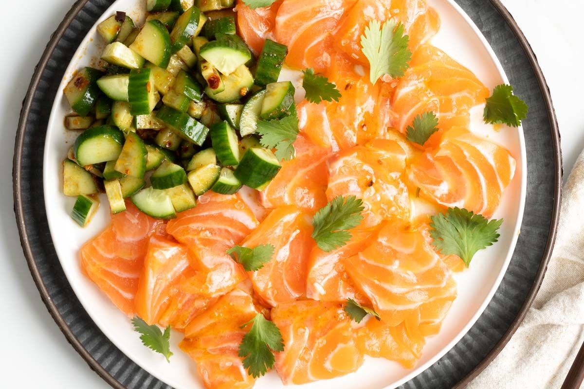 Sliced raw salmon arranged on a plate with chopped cucumbers and garnished with fresh cilantro leaves.