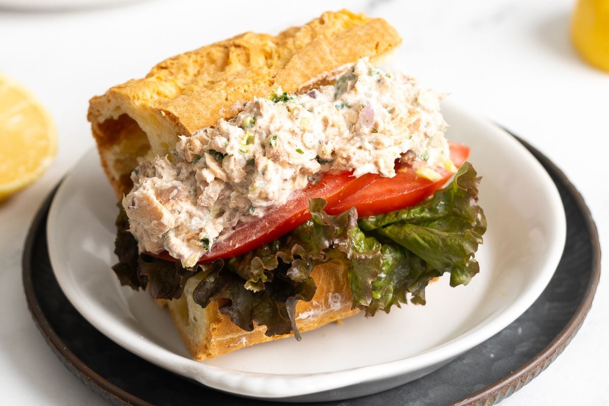 A sandwich on a white plate, filled with tuna salad, sliced tomato, and leafy lettuce in a crusty bread roll.