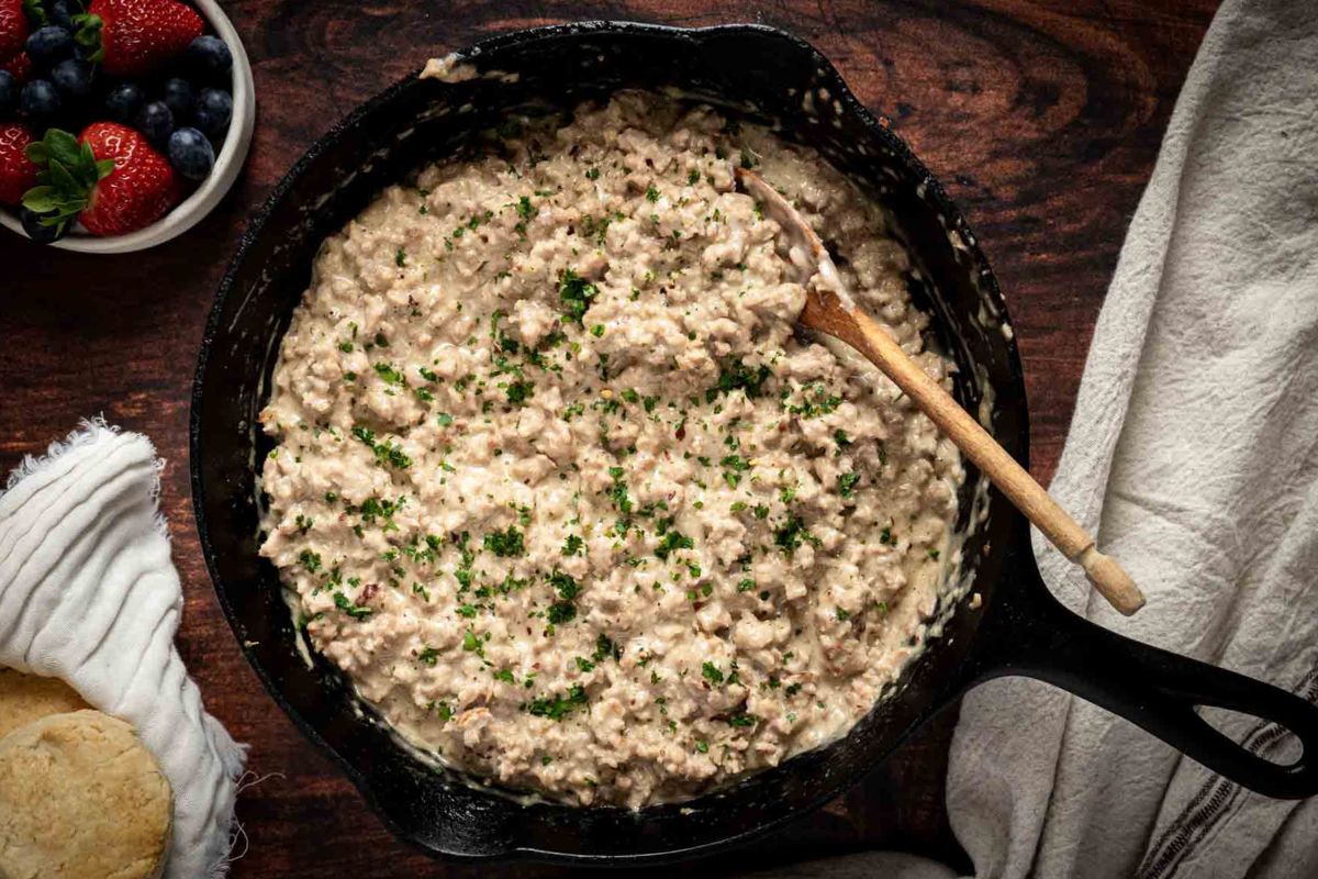 A cast iron skillet filled with creamy sausage gravy, garnished with chopped herbs, sits on a wooden surface with a wooden spoon and a biscuit nearby.