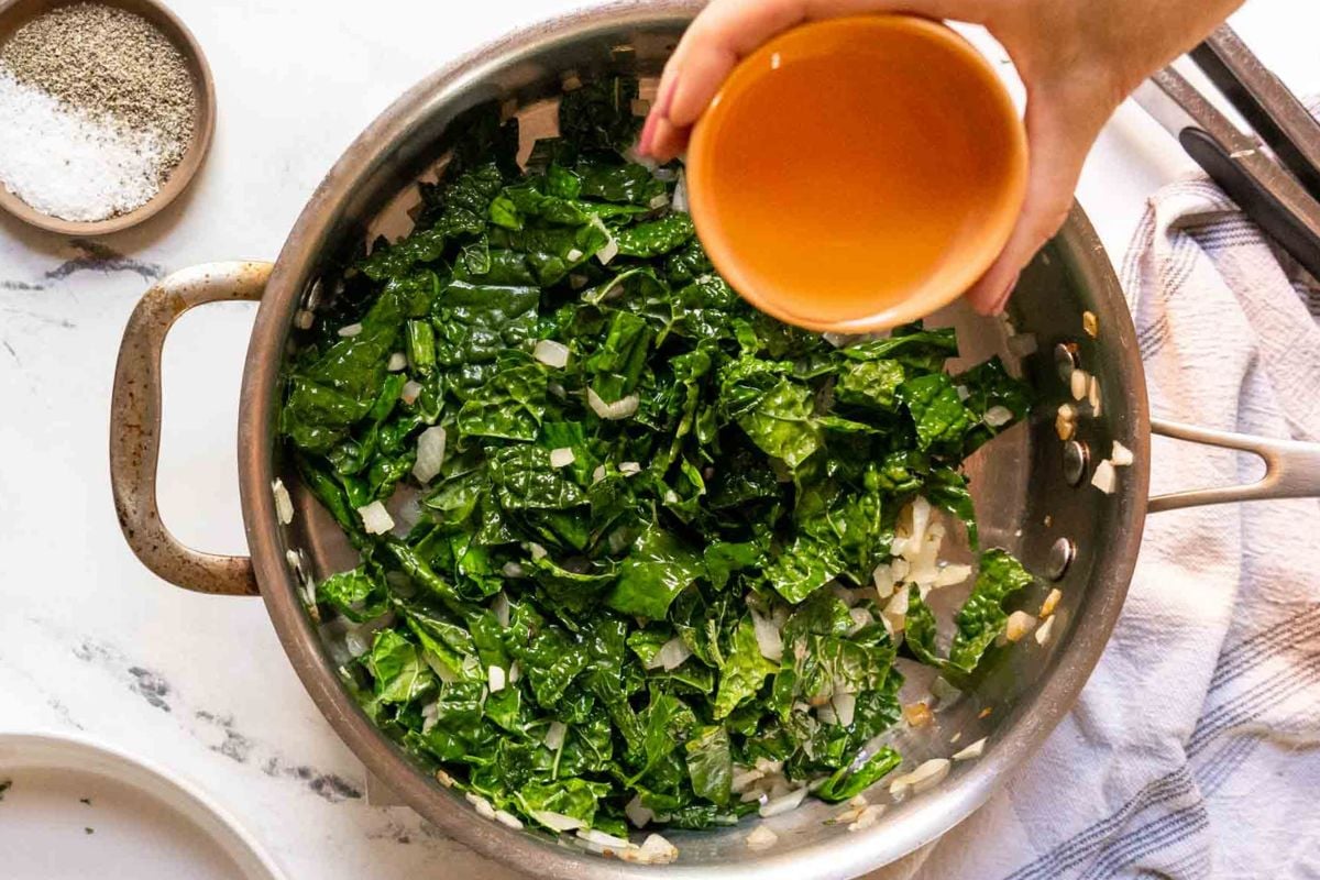 A hand pours liquid from a small orange bowl into a pan of chopped greens and onions cooking on a stovetop.